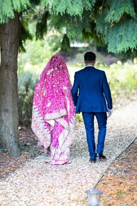 A photograph of a couple walking along a tree lined gravel path, he is dressed in a western blue suit, his partner is dressed in a beautiful pink traditional lengha.